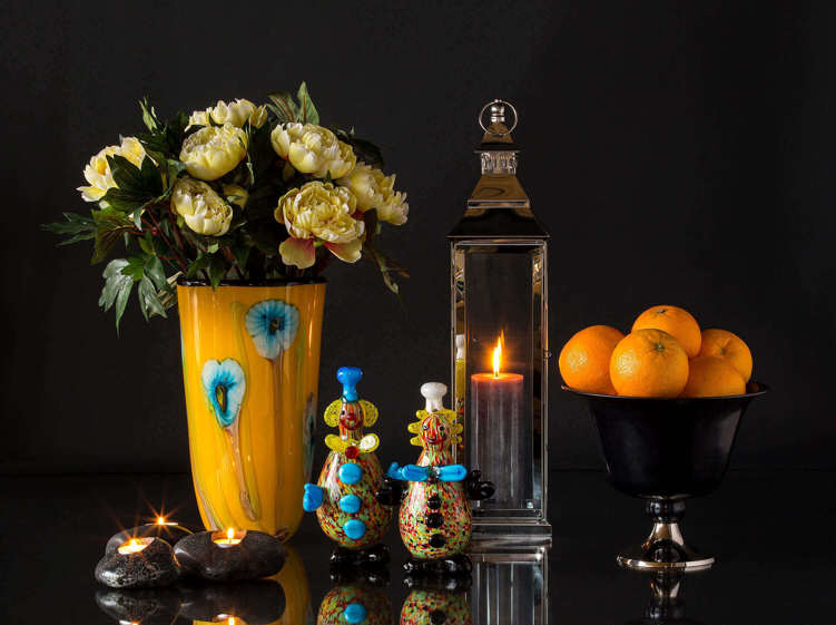 To klovnefigurer i glas foran gul glasvase, lanterne og frugtskål