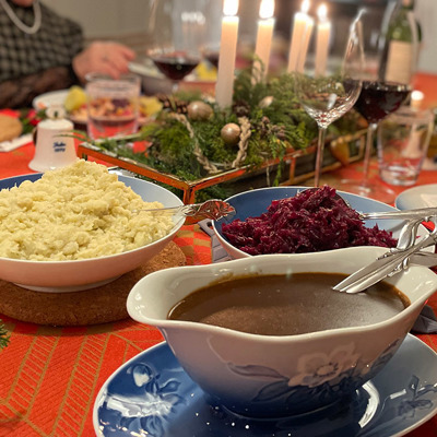 Juleaften med klassisk julemad og bord dækket med Bing & Grøndahl Julerose stel. Serveringsfade med rødkål, kartofler og sauce står på rød dug omgivet af julepynt og stearinlys.