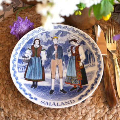 Ravn platte med motiv af folkedragter fra Småland i Sverige, brugt som tallerken i en borddækning med fletbrik, blomster og gyldent bestik.