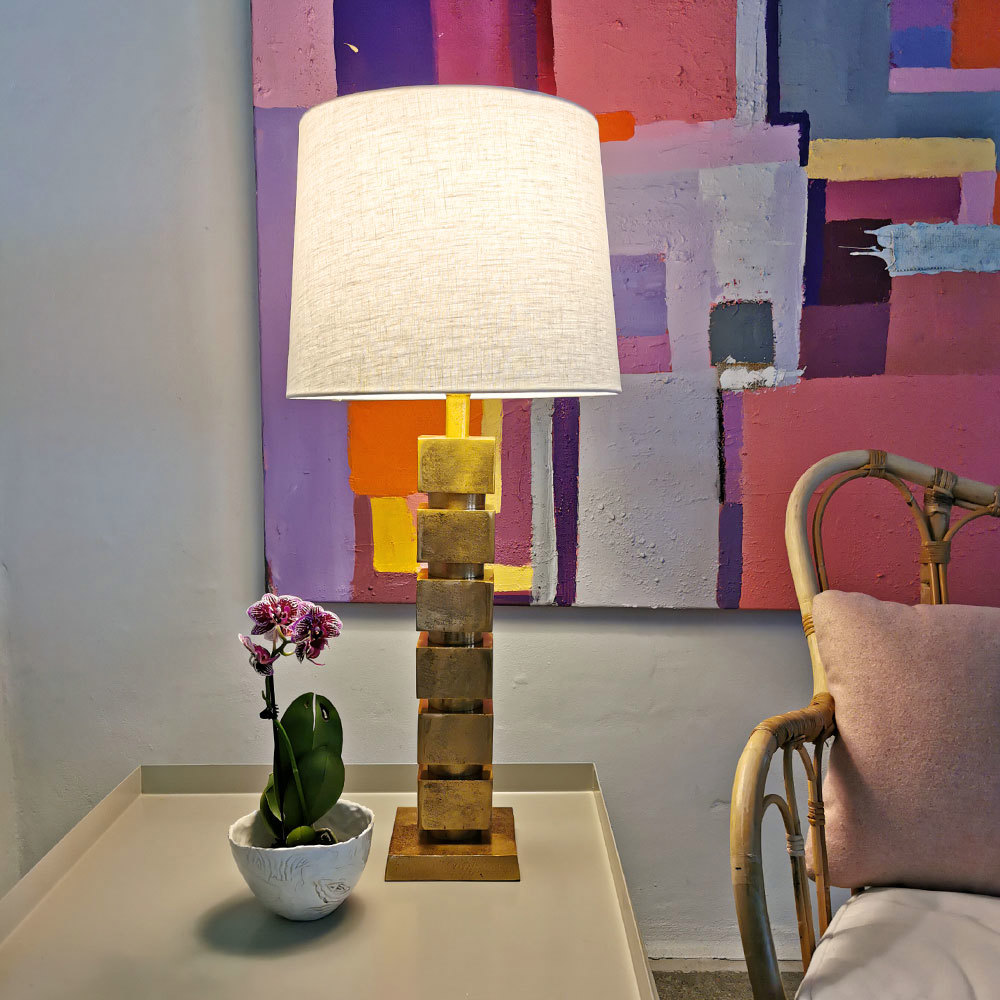 Cylinderformet lampeskærm i lys hør (farve 1000) med naturlig struktur, monteret på bordlampe med gylden fod på et lyst sidebord.