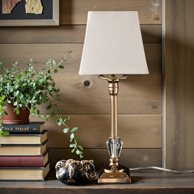 Klassisk bordlampe med gyldent finish, stor krystaldetalje og firkantet lampeskærm placeret på sidebord i hyggelig indretning.