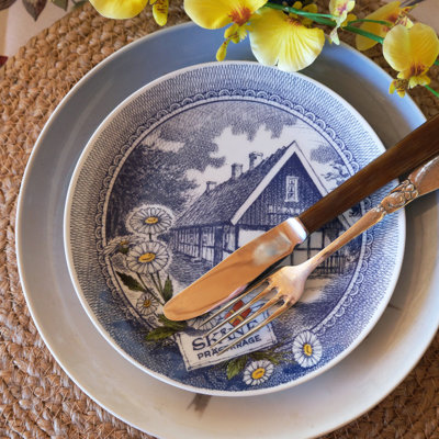 Borddækning med Ravn landskabsplatte brugt som forretstallerken ovenpå et blåt stel. Fletbrik og blomster i gule nuancer fuldender det personlige udtryk.