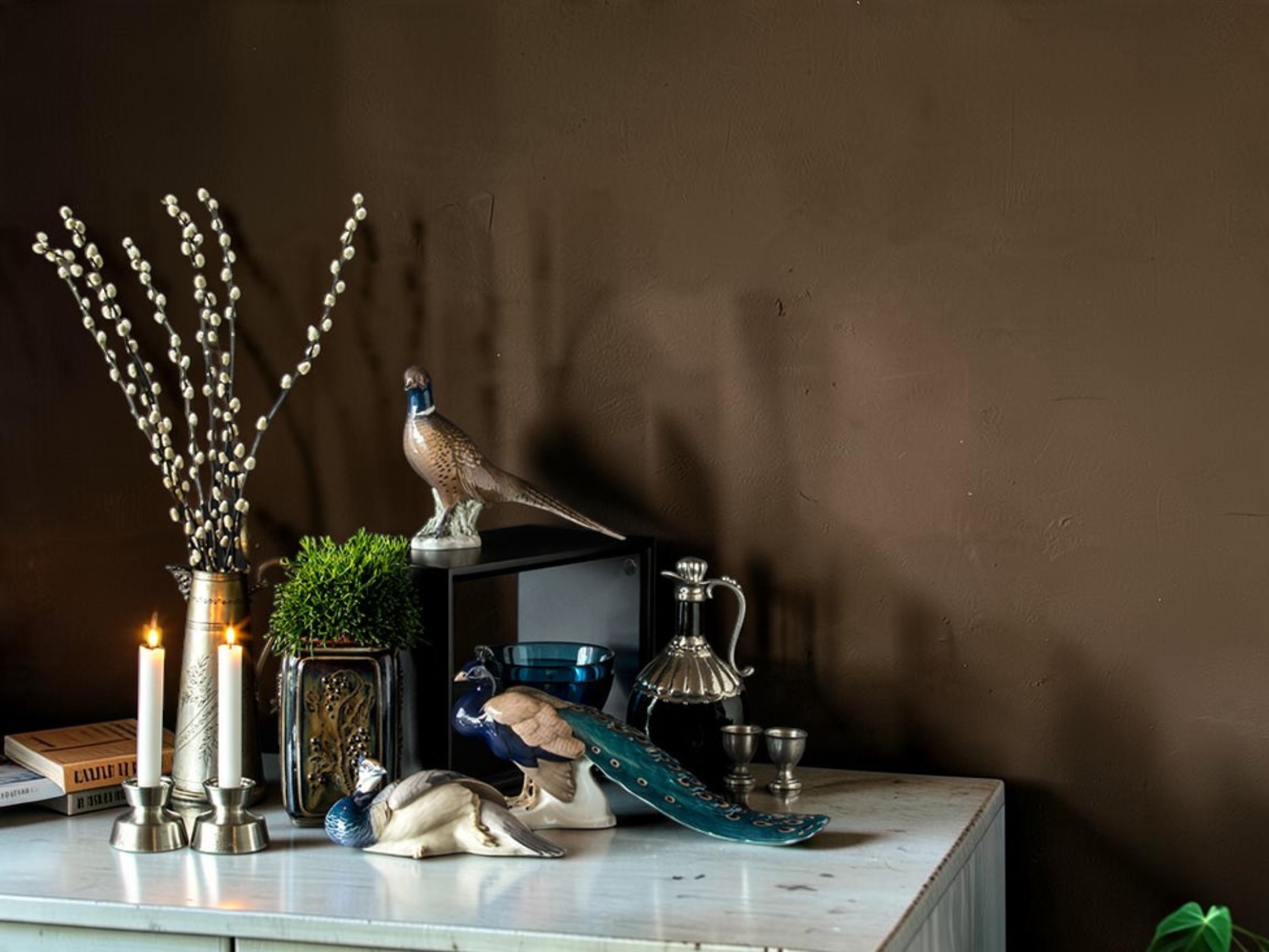 Dekorative porcelænsfugle fra Bing & Grøndahl opstillet med lysestager og grene foran en mørk væg.