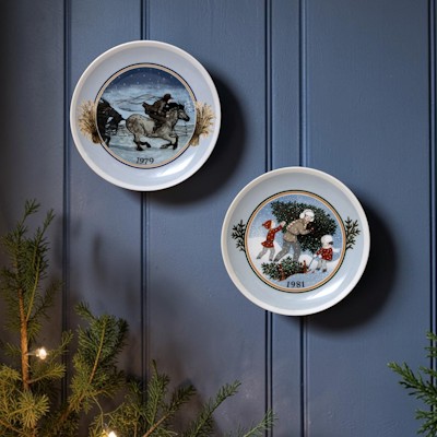To Tettau juleplatter med vintermotiver hænger på en mørk blå væg over gran og varme julelys.