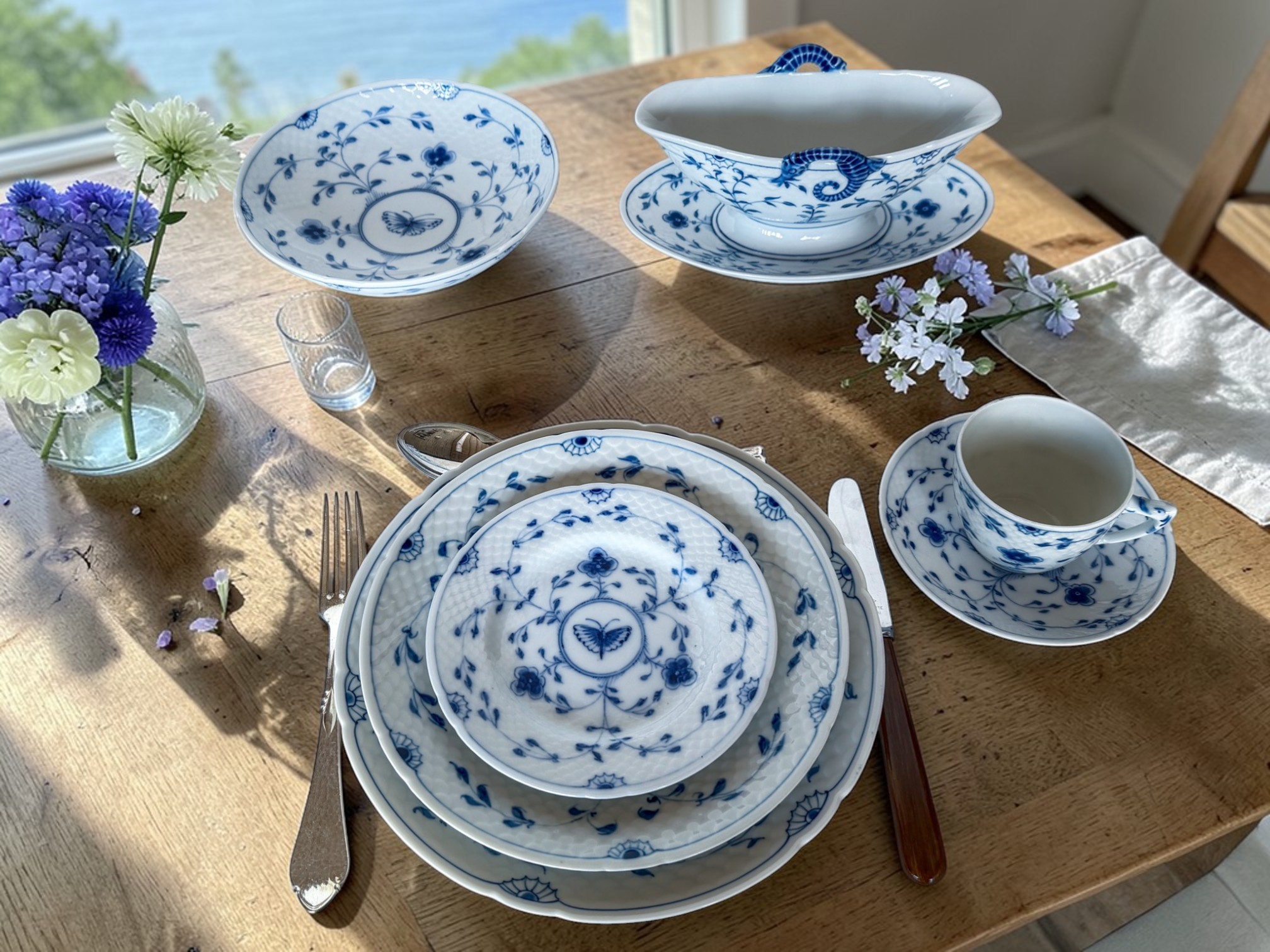 Bing & Groendahl Sommerfuglestel i blå og hvid porcelæn opdækket med middagstallerken, dyb tallerken, skål, kaffekop og sovseskål på træbord med blomster og udsigt til vand